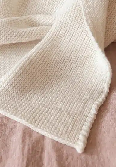 Cream-colored knitted blanket with a waffle-like texture draped over a pink wooden surface