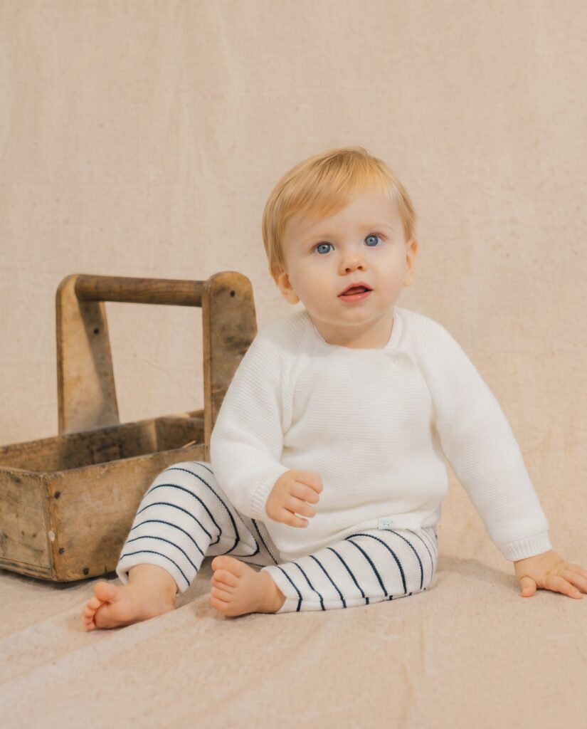 Baby with blue eyes sits on the floor beside a rustic wooden chair, wearing a white sweater and striped leggings.