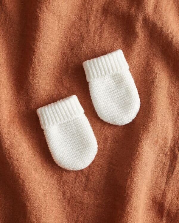 Two white knitted baby mittens resting on an orange fabric background.