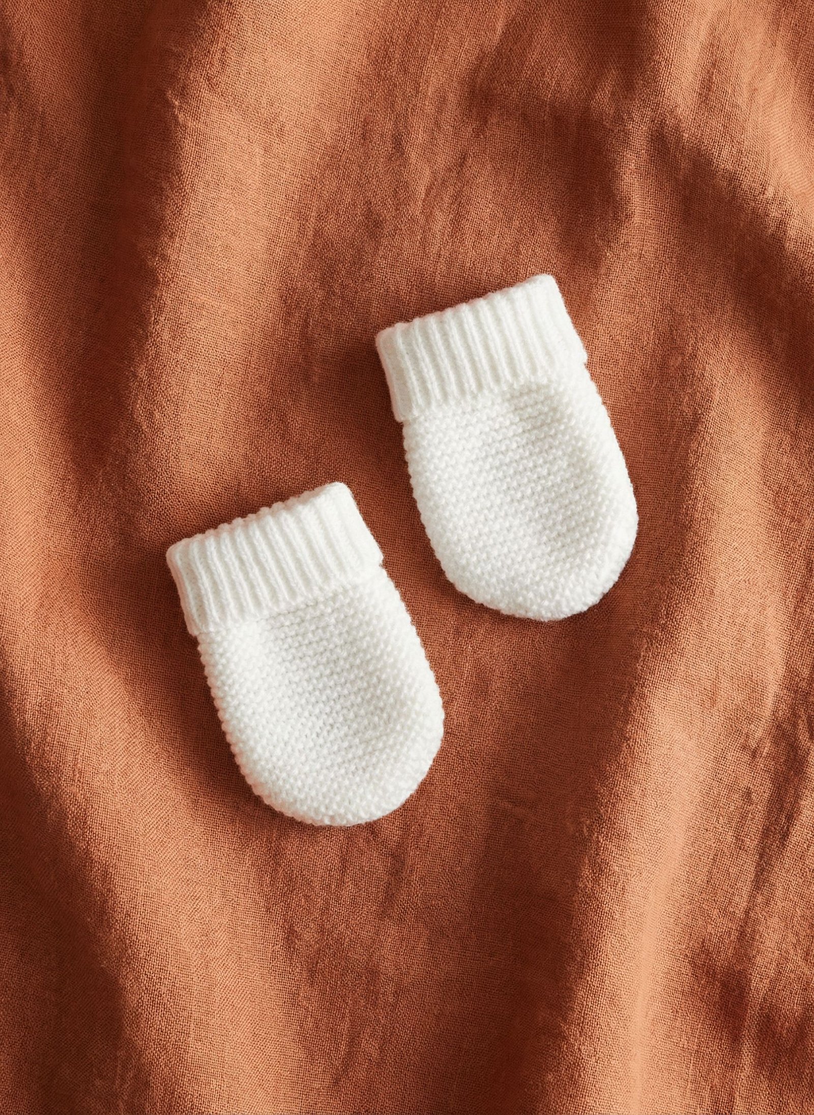 Two white knitted baby mittens resting on an orange fabric background.