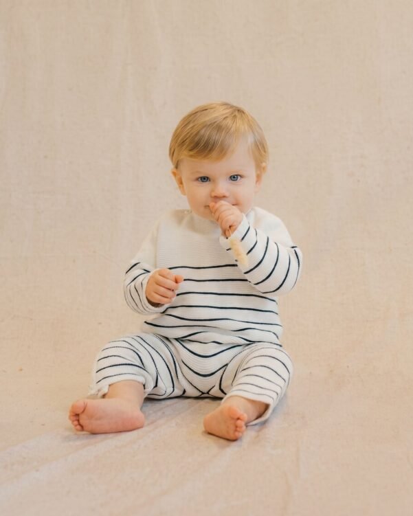 Baby sits on a light beige backdrop in a white/navy striped outfit, sucking their thumb and looking at the camera.