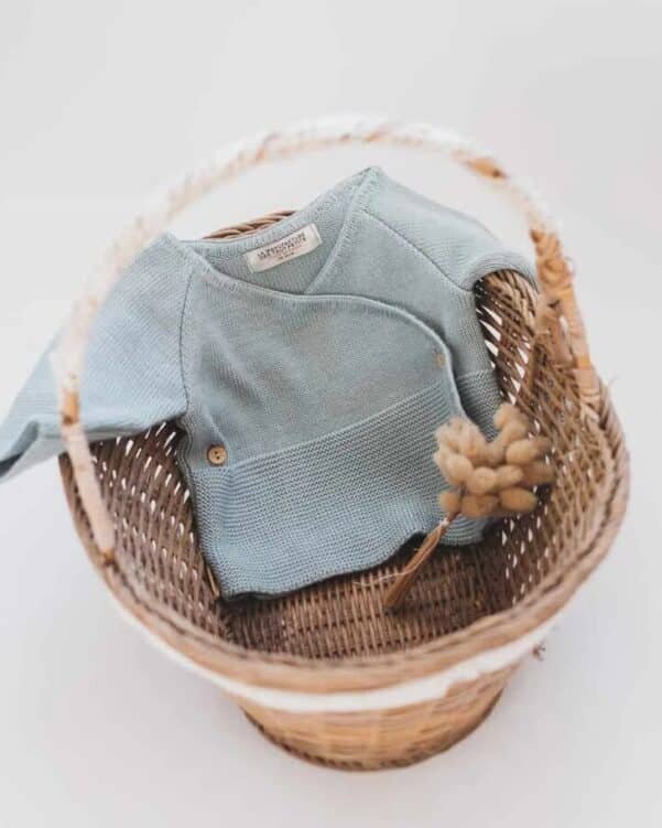 Blue knitted baby onesie folded in a wicker basket with a dried grass stem for a soft, cozy display.