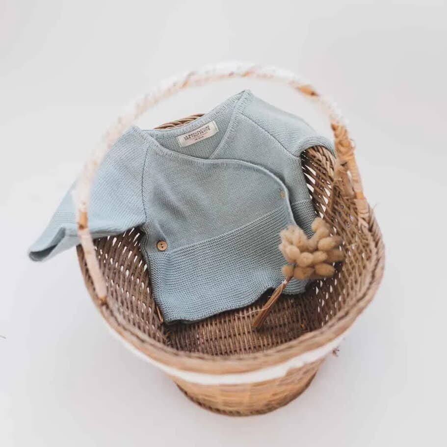 Blue knitted baby onesie folded in a wicker basket with a dried grass stem for a soft, cozy display.